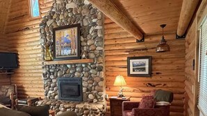 A cozy log cabin interior with a stone fireplace, a sofa, a chair, and a lamp. - Sagebrush Meadows+Hotub+WiFi+20minsYellowstone (Island Park)