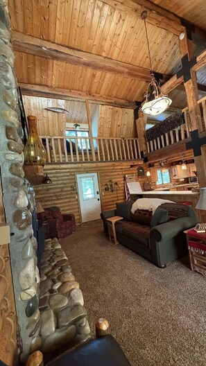 A log cabin interior with a stone wall, a wooden ceiling, a sofa, a dining area, and a staircase. - Sagebrush Meadows+Hotub+WiFi+20minsYellowstone (Island Park)