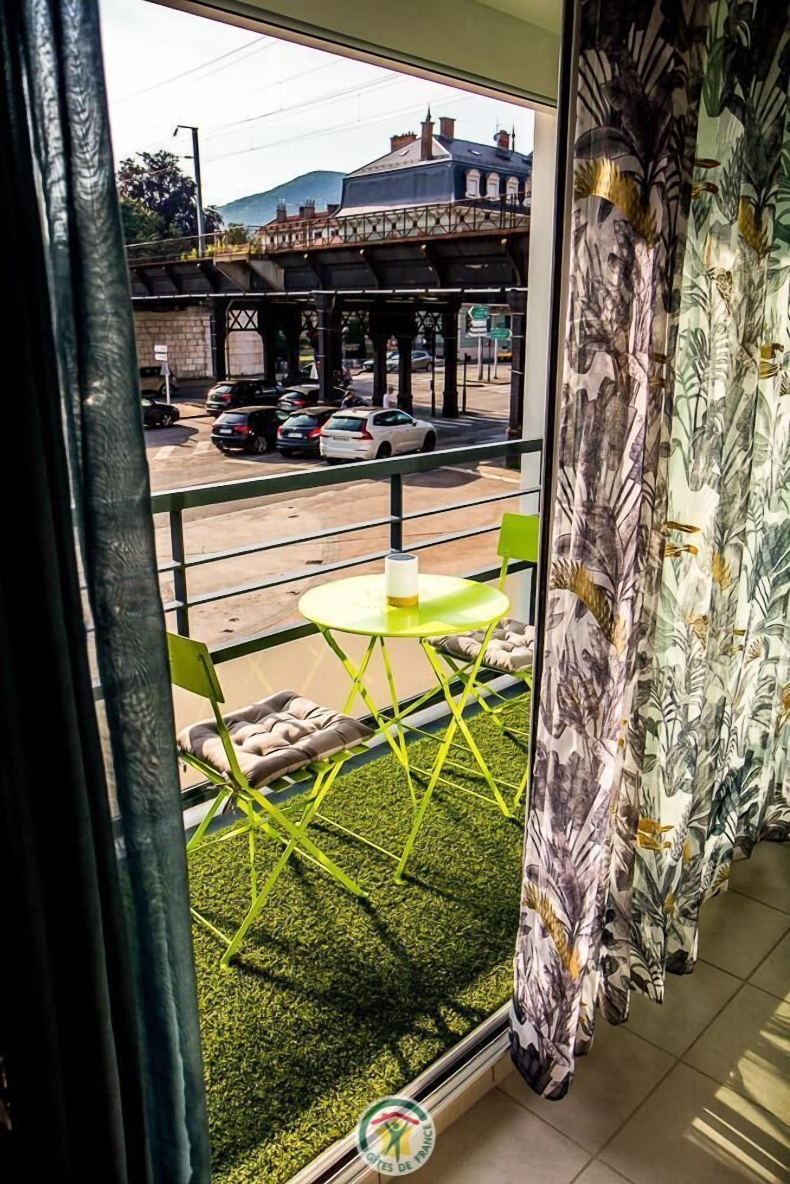 Un balcon avec une chaise verte et une petite table, donnant sur une rue où circulent des voitures et sur un pont.