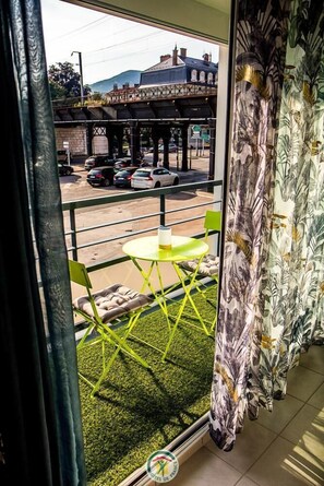 Ein Balkon mit einem grünen Stuhl und einem kleinen Tisch, mit Blick auf eine Straße mit Autos und einer Brücke.