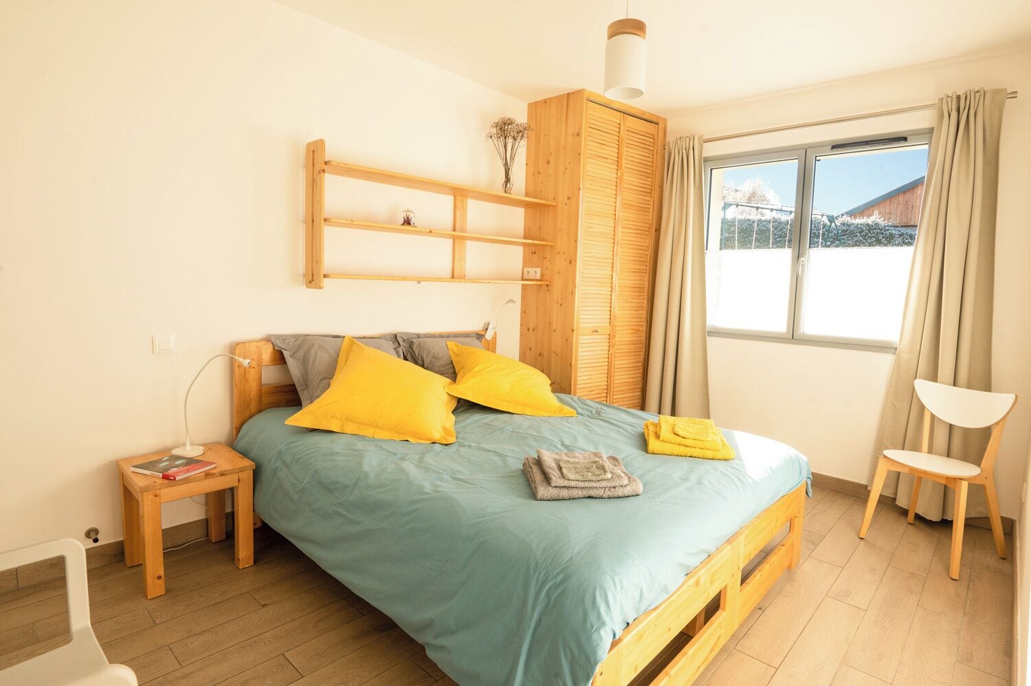 A bedroom with a bed, wooden headboard, a bedside table with a lamp, a chair, and a window with curtains.