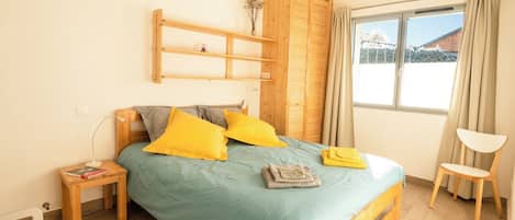 A bedroom with a bed, wooden headboard, a bedside table with a lamp, a chair, and a window with curtains.