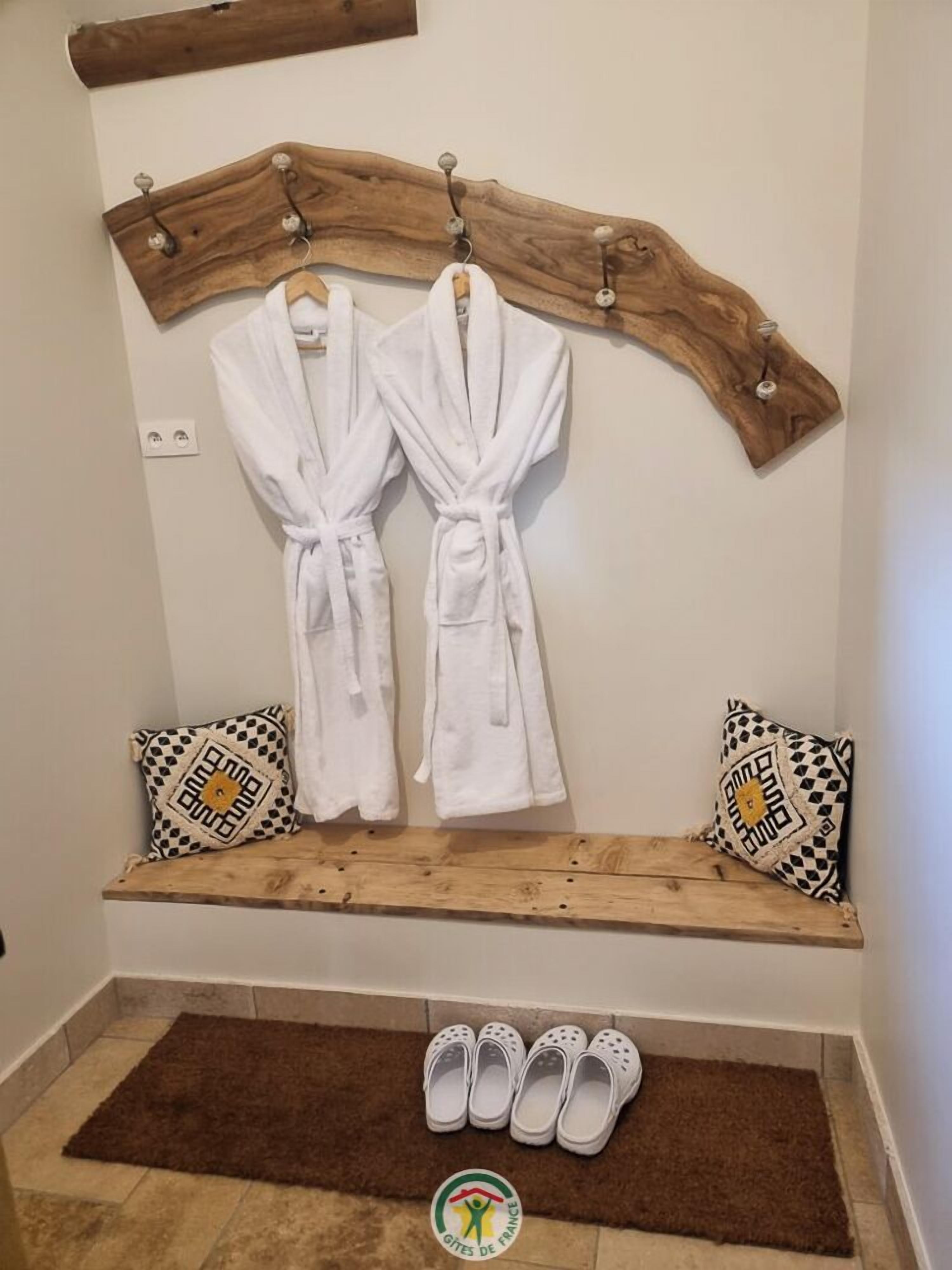 Un baño con un estante de madera que sostiene dos albornoces blancos, un banco con cojines estampados y un par de zapatillas blancas sobre un tapete marrón.