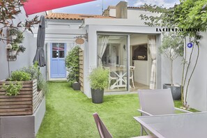 Ein kleiner Innenhof mit einer Holzbank, einem Tisch und Stühlen. Ein blauer und eine Glastür führen in einen Raum.