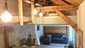 A loft-style living space with exposed wooden beams, a dining area with a wooden table set for four, and a blue sofa against a textured wall. - Le Nid in Chartreuse (SAINT PIERRE DE CHARTREUSE)