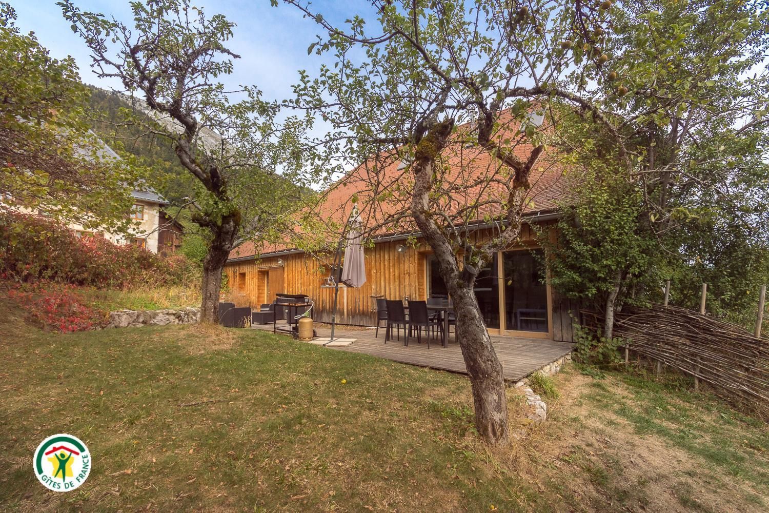Une maison en bois avec une terrasse, du mobilier d’extérieur et un mur en pierre.