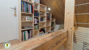 A wooden bookshelf with books and a lamp, a lofted sleeping area with a bed, and a wooden railing. - Le Nid in Chartreuse (SAINT PIERRE DE CHARTREUSE)