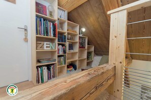 A wooden bookshelf with books and a lamp, a lofted sleeping area with a bed, and a wooden railing. - Le Nid in Chartreuse (SAINT PIERRE DE CHARTREUSE)