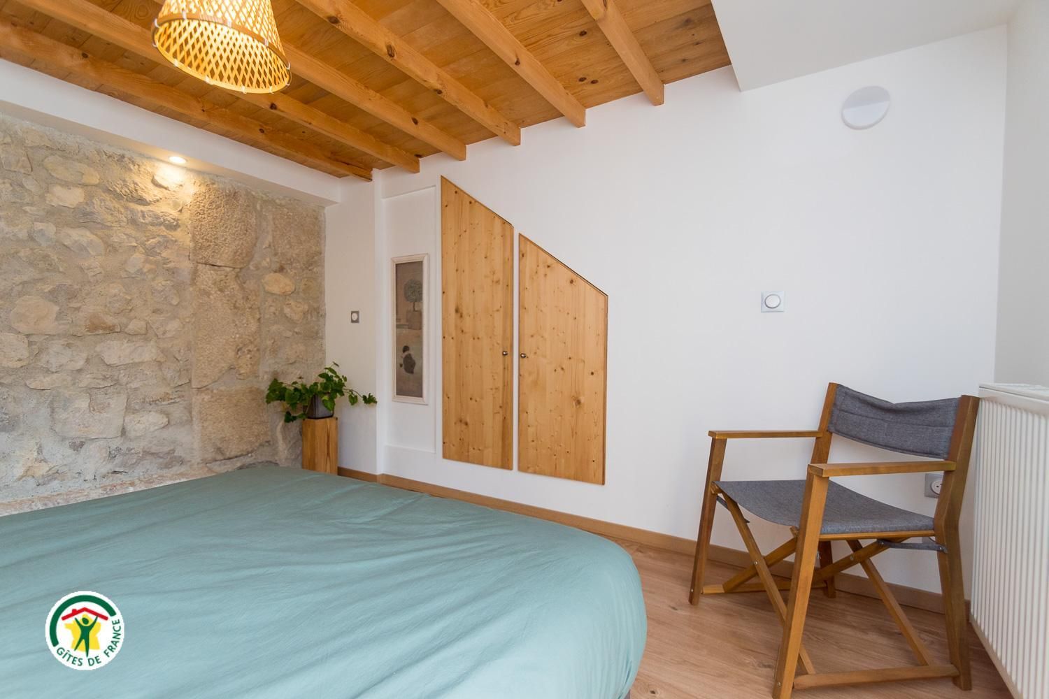 A room with a bed, a chair, a stone wall, wooden ceiling beams, and a hanging light fixture.