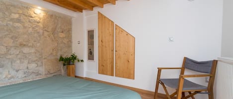 Ein Zimmer mit einem Bett, einem Sessel, einer Steinwand, Holzbalken an der Decke und einer Hängelampe.