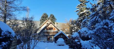 Una cabaña de madera con techo pronunciado, rodeada de árboles cubiertos de nieve y un cielo azul despejado.