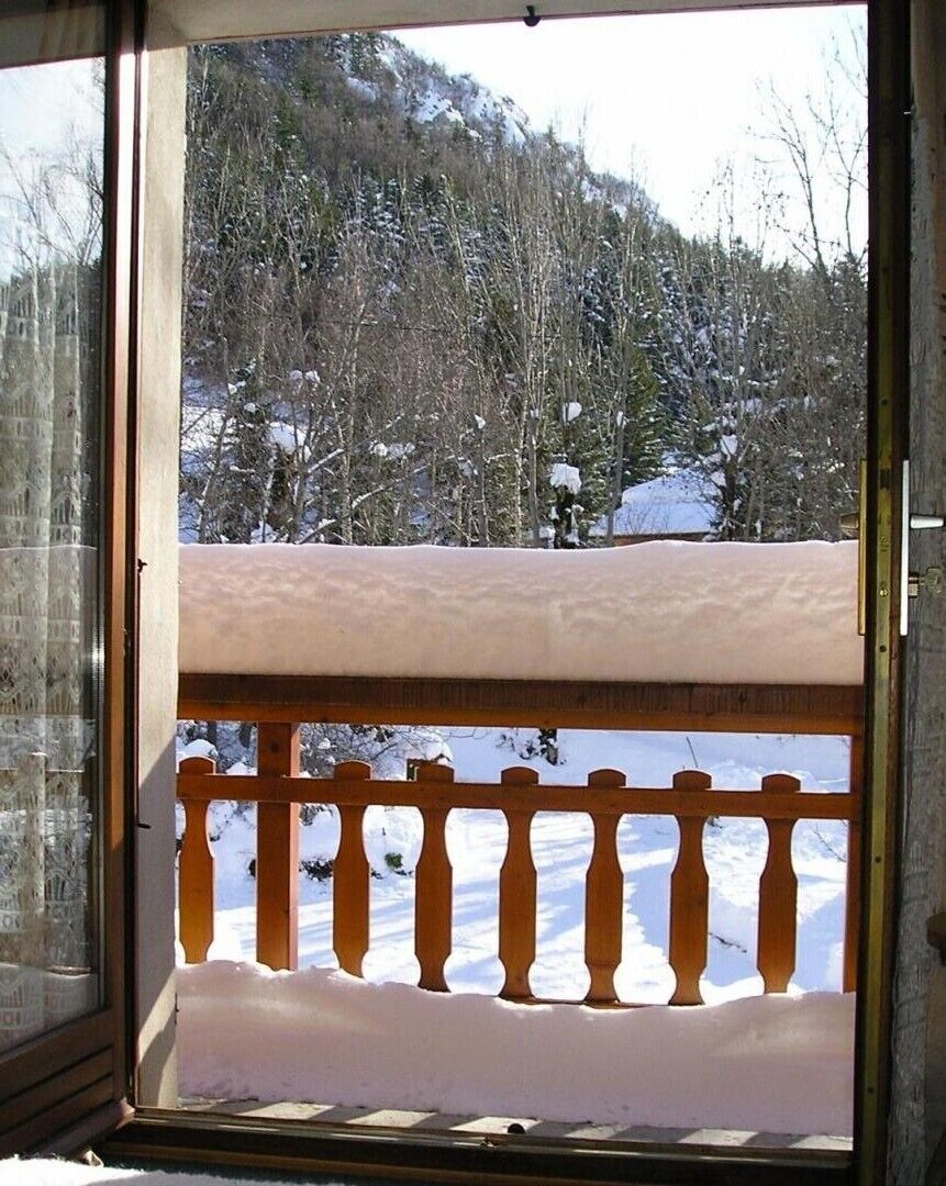 A view from a balcony with a wooden railing, overlooking a snowy landscape with trees and a mountain in the background.