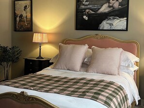 Ein ordentlich bezogenes Bett mit kariertem Überwurf, einer Nachttischlampe und zwei Kissen. An der Wand über dem Bett hängen gerahmte Kunstwerke.