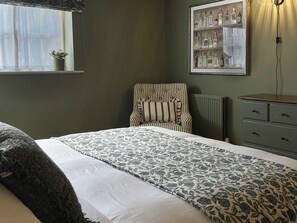 Ein Schlafzimmer mit einem Bett, einem Sessel, einem Schminktisch, einer Heizung und einem an der Wand befestigten Regal mit Flaschen.