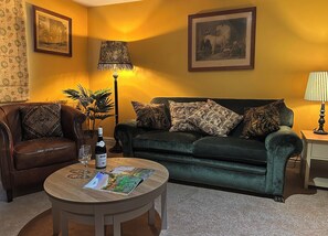 Ein Wohnzimmer mit einem Ledersofa, einem grünen Sofa mit gemusterten Kissen, einem runden Couchtisch mit Weinflasche und Glas sowie zwei Wandleuchten.