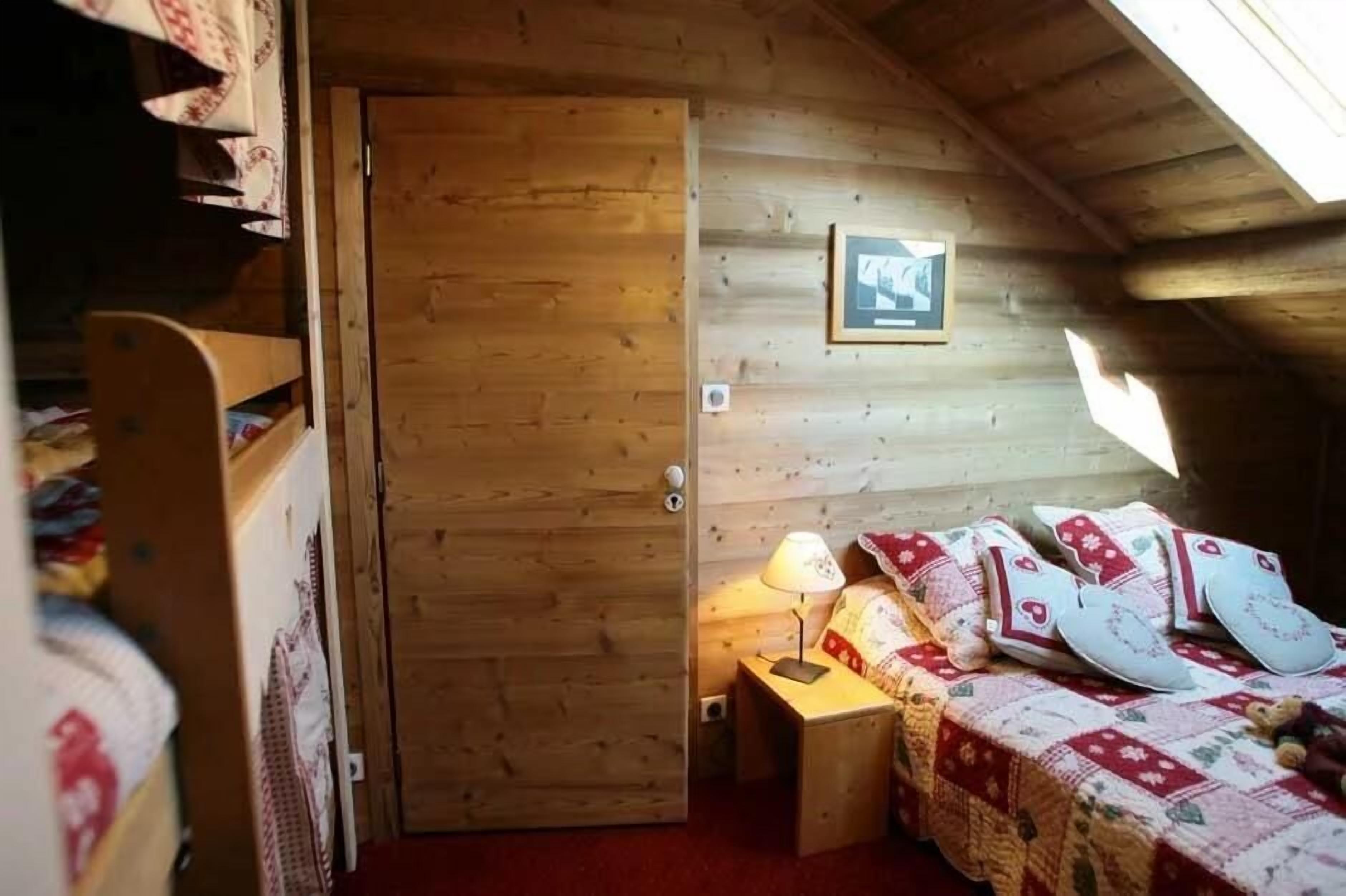 A cozy room with a wooden door, a bed with patterned bedding, a small table with a lamp, and a framed picture on the wall.