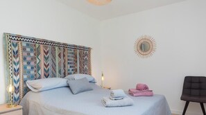 Ein ordentlich bezogenes Bett mit Kissen, ein an der Wand befestigter Spiegel und ein Sessel in einem Raum mit gemusterten Vorhängen.