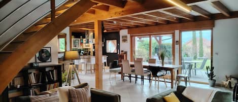 A spacious living area with a high ceiling, wooden beams, a dining area with chairs, and a view of the outdoors through large windows.