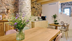 Una mesa de madera para comer con un florero, una pared de piedra, una zona de cocina con un fregadero y una computadora, y una escalera que lleva a un nivel superior.