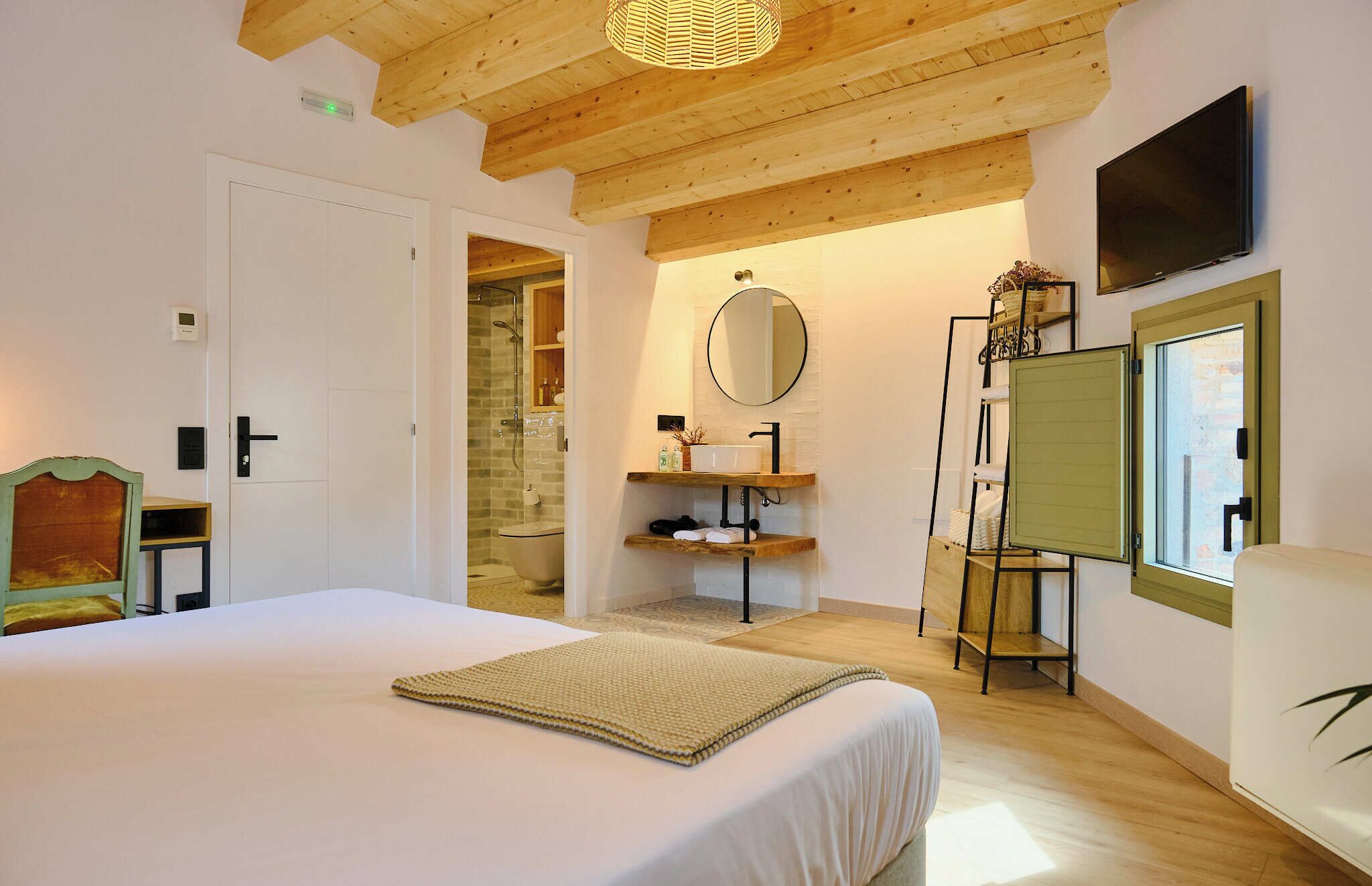 Un dormitorio con cama, techo de madera, baño con lavabo y espejo, y una ventana.