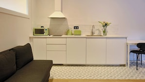 Cocina compacta con microondas, hervidor, tostadora y lavabo. Hay una pequeña mesa de comedor con una silla y un sofá en la sala.