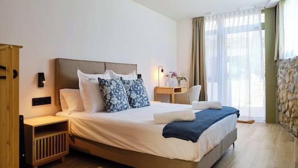 Habitación de hotel con una cama grande, mesitas de noche de madera, una mesita con lámpara y vistas al exterior a través de cortinas translúcidas.