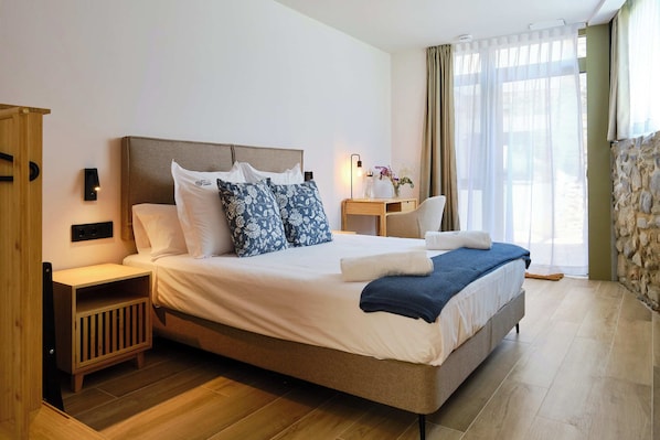 A hotel room with a large bed, wooden nightstands, a bedside table with a lamp, and a view of the outdoors through sheer curtains.