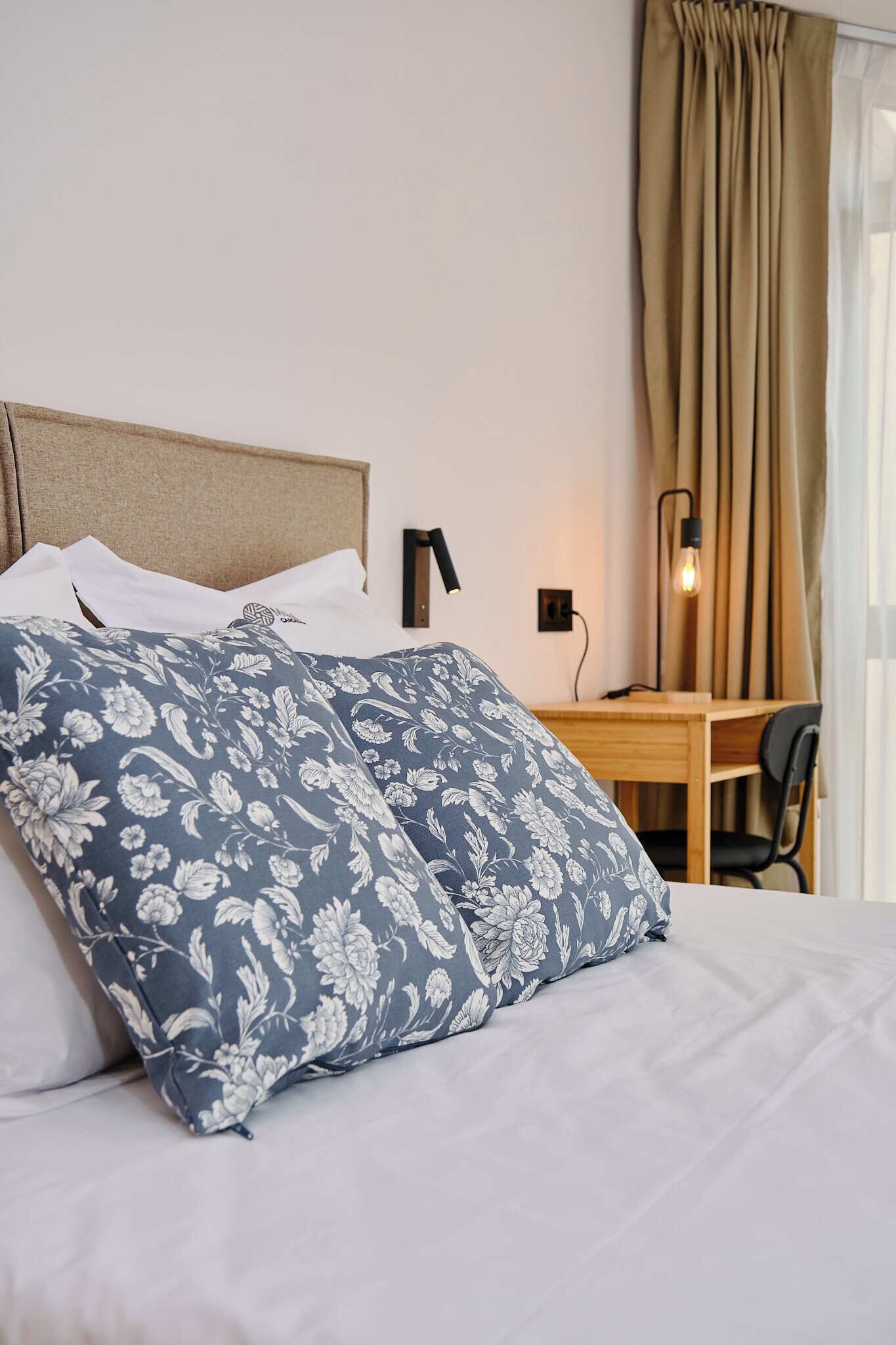 Habitación de hotel con cama, escritorio, silla, lámpara y ventana con cortinas.