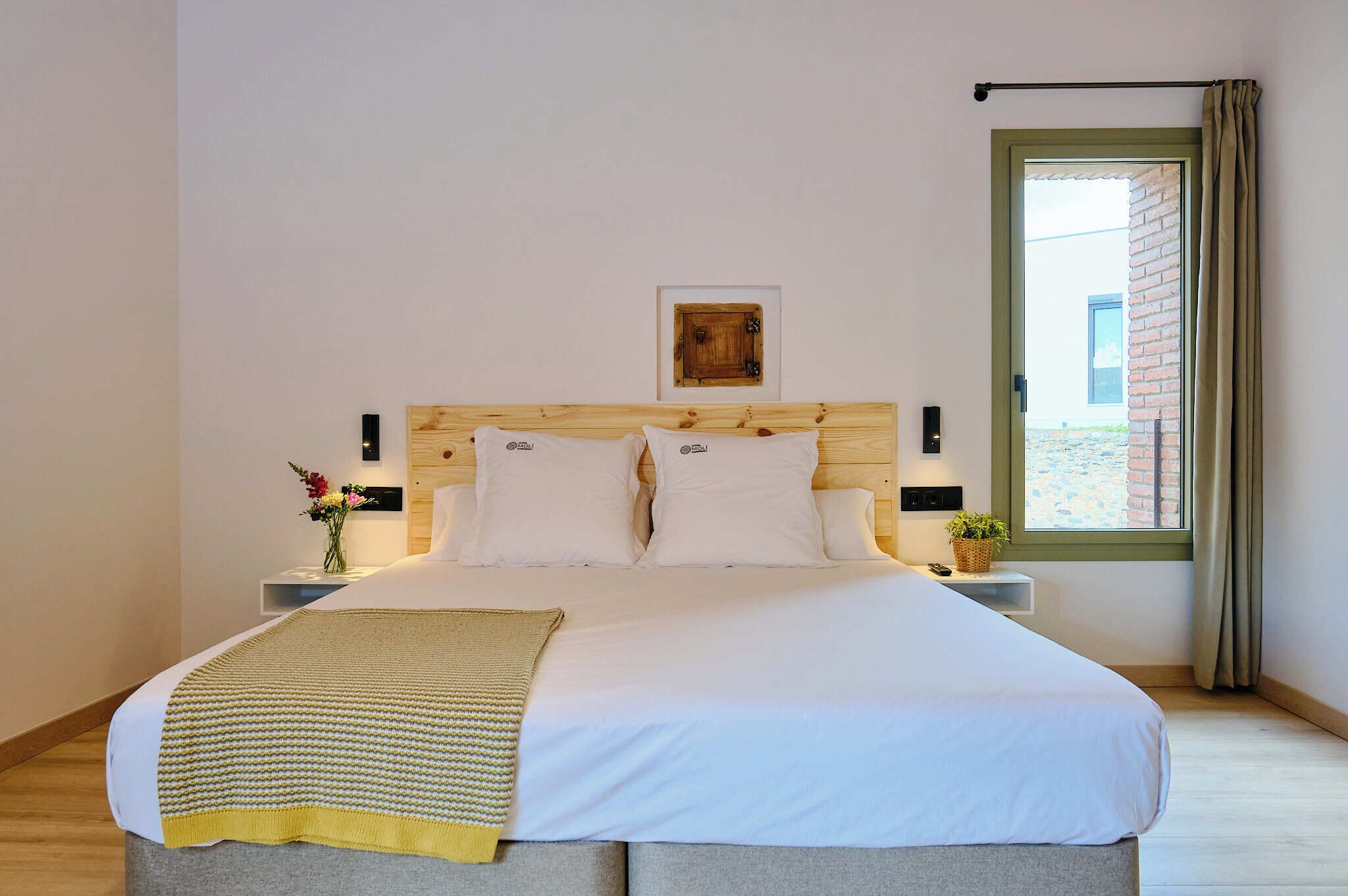 A neatly made bed with a wooden headboard, white bedding, and a yellow throw blanket. There is a small bedside table with a vase of flowers and a wall-mounted light. A window with curtains is visible, and a framed picture hangs above the bed.