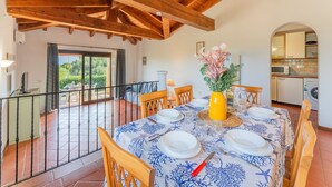 Un comedor con una mesa preparada para cuatro, un jarrón amarillo con flores, y vistas de la cocina y el patio exterior.