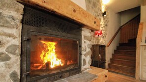 Una chimenea de piedra con fuego visible, una viga de madera encima y una escalera al fondo.