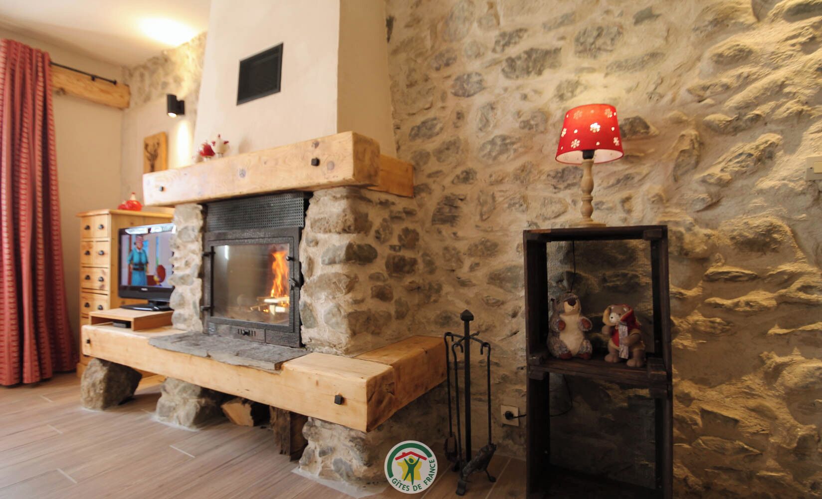 A stone fireplace with a television on a wooden stand, a red lamp, and a shelf with stuffed animals.