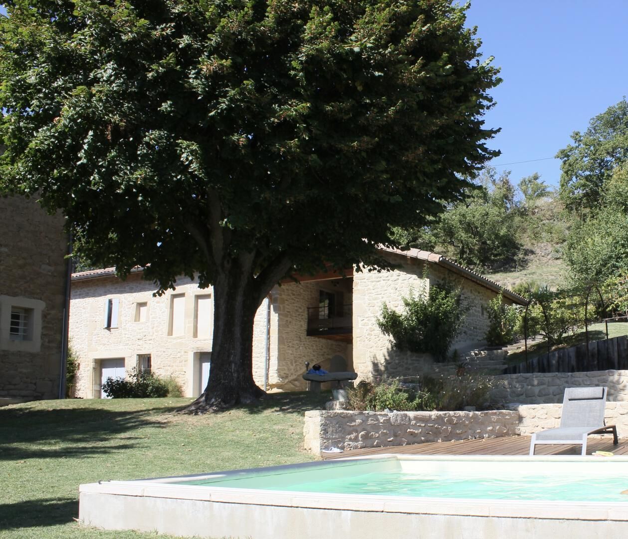 Un bâtiment en pierre avec une piscine, un grand arbre et une terrasse en bois.