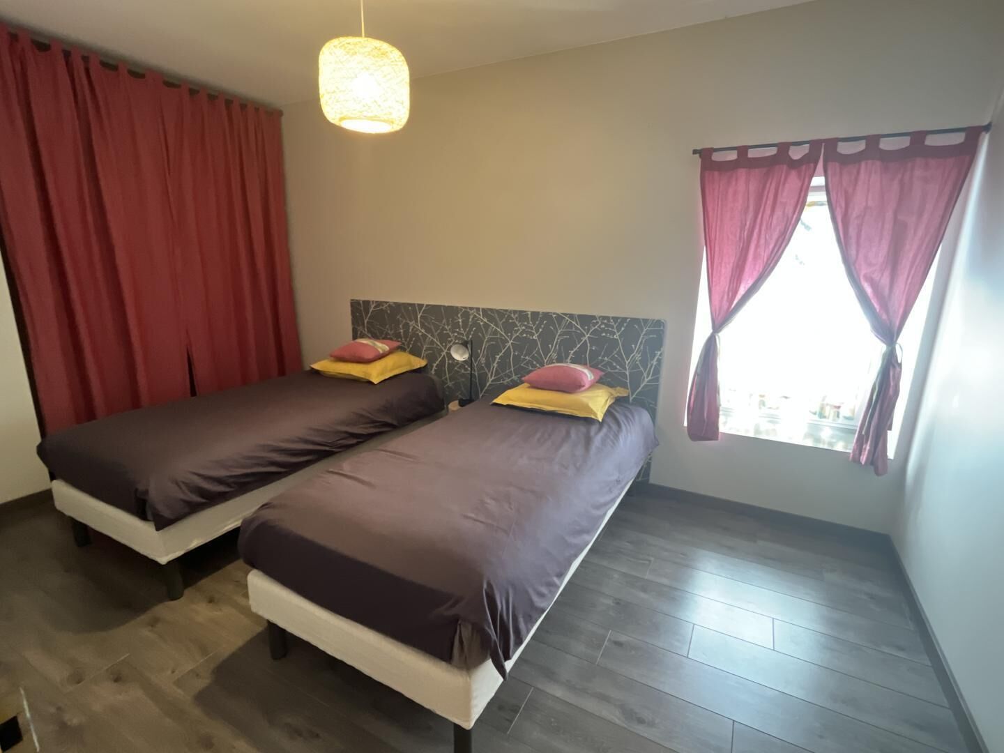 A double bed with a headboard, two pillows, and a hanging light fixture in a room with wooden flooring and a window with curtains.