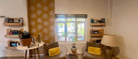 Sala de estar com dois sofás, uma mesa de centro e uma luminária. Estantes exibem livros e objetos decorativos. Uma janela revela vista para o exterior.