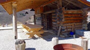 Une cabane rustique en rondins de bois, dotée d’une terrasse en bois et d’un foyer extérieur en métal.