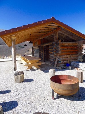 Une cabane rustique en rondins de bois, dotée d’une terrasse en bois et d’un foyer extérieur en métal.