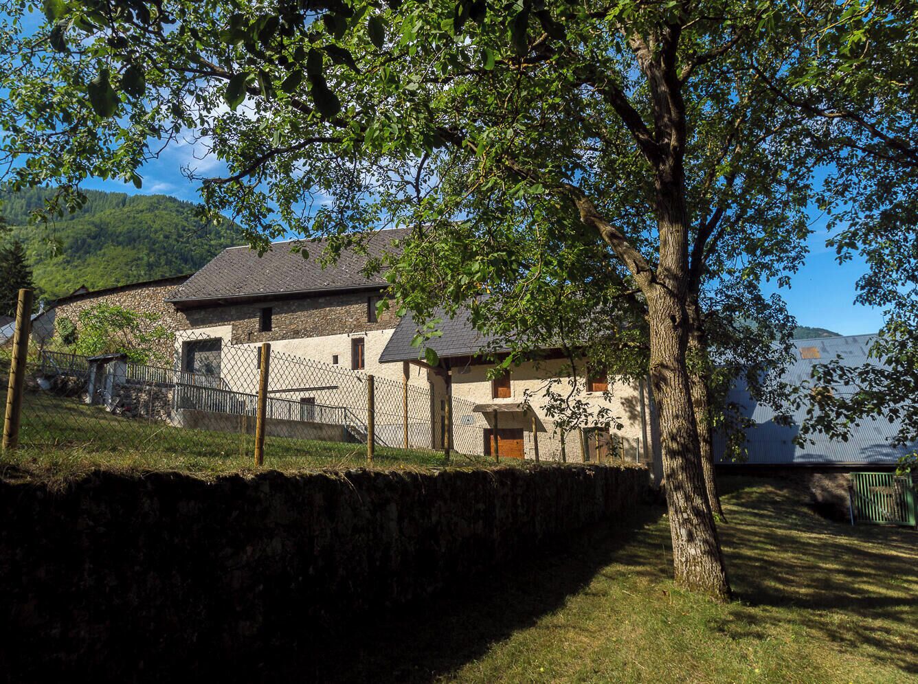 A stone building with a sloped roof, surrounded by a stone wall and a fenced area.