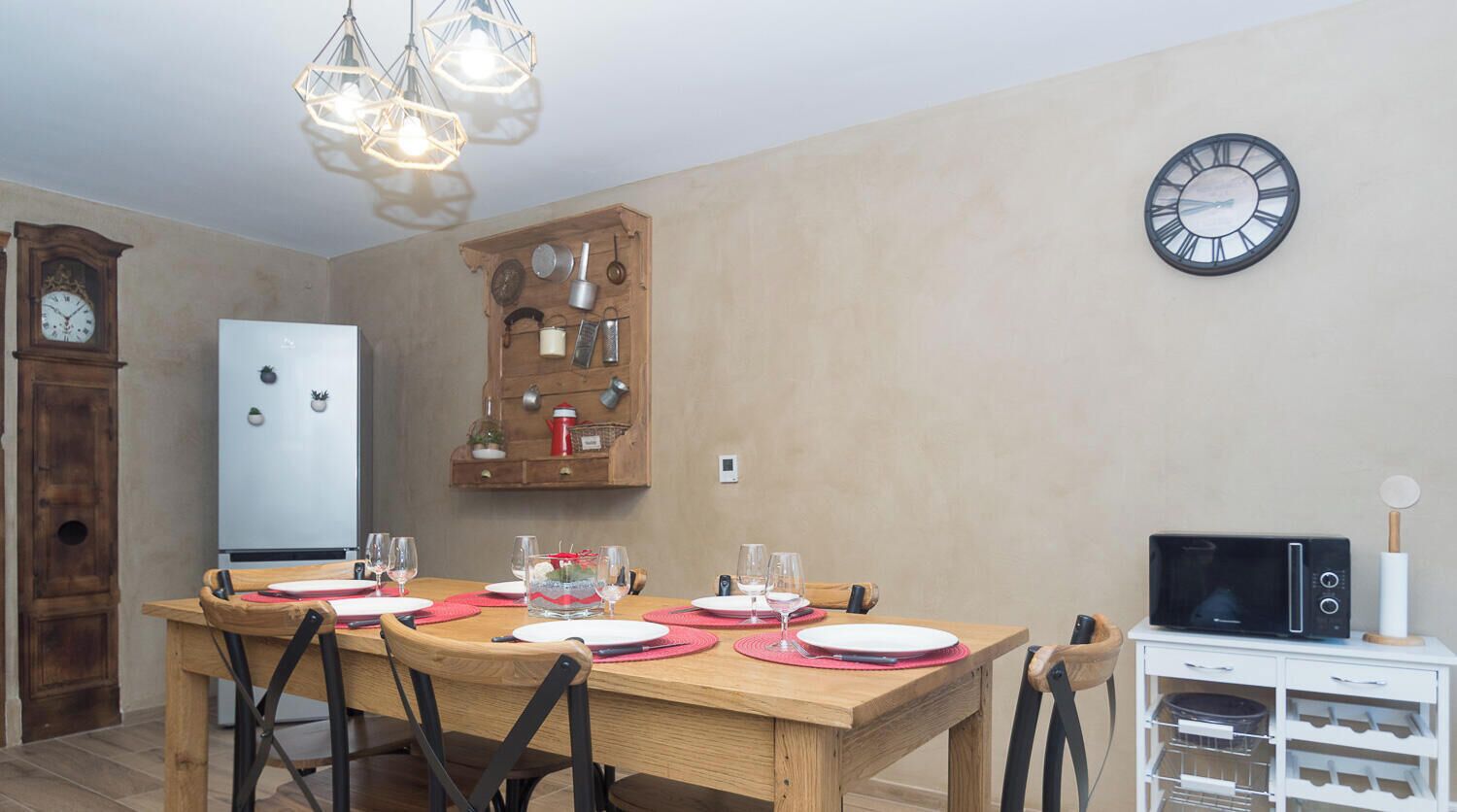 A dining area with a wooden table set for four, surrounded by chairs. A wall clock and a microwave are visible in the background.