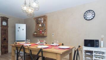 A dining area with a wooden table set for four, surrounded by chairs. A wall clock and a microwave are visible in the background.