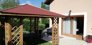 Ein überdachter Außenbereich mit Tisch und Stühlen, einer Holz-Pergola und einer Schiebetür zu einer Terrasse.
