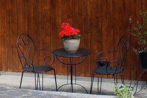 Ein kleiner Sitzbereich im Freien mit einem runden Tisch und zwei Stühlen, einem Blumentopf mit roten Blüten und einem Holzzaun im Hintergrund.