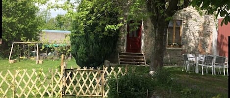 Un bâtiment en pierre avec une porte rouge, entouré d’une clôture en bois, et un arbre au premier plan.