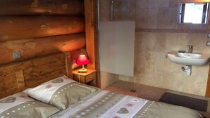 A wooden cabin-style room with a bed, a nightstand with a lamp, a bathroom with a sink and mirror. - CHANTELOUVE / LE BECAN (CHANTEPERIER)