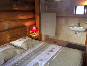 Ein holzgetäfeltes Zimmer im Blockhausstil mit Bett, Nachttisch mit Lampe und einem Badezimmer mit Waschbecken und Spiegel.