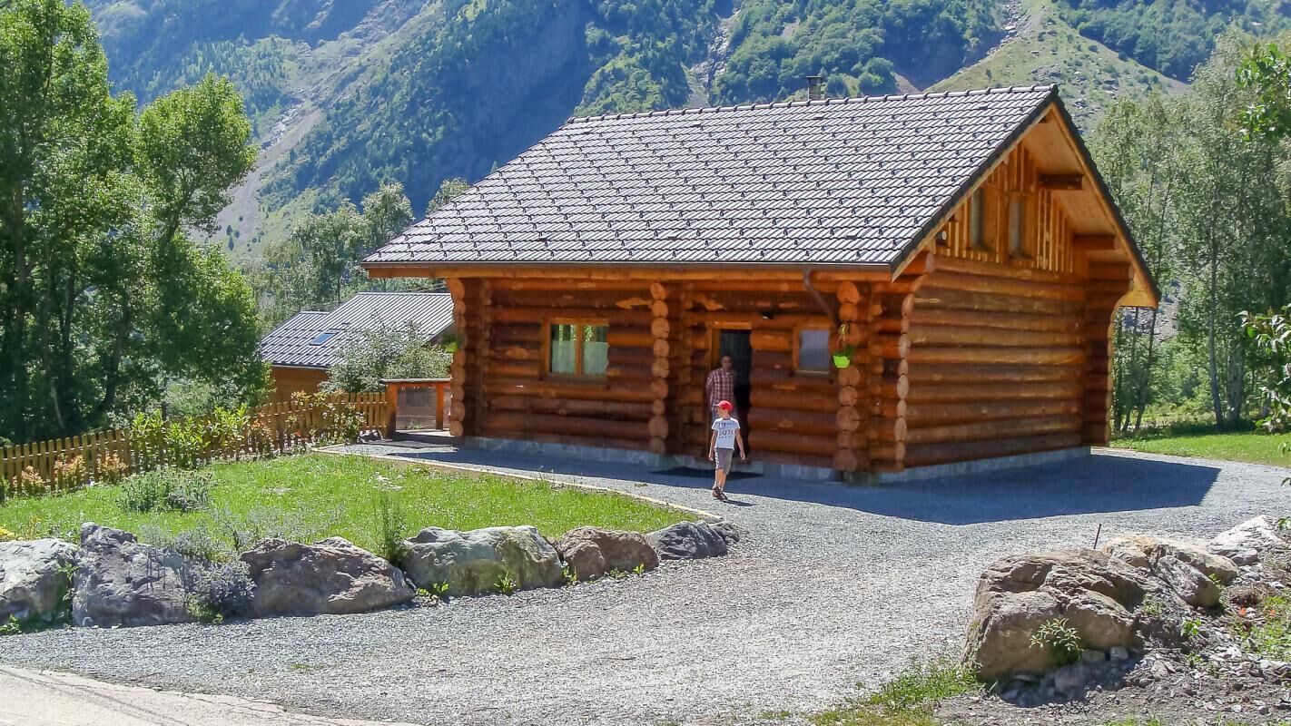 A wooden log cabin with a sloped roof, surrounded by a gravel driveway and a wooden fence.