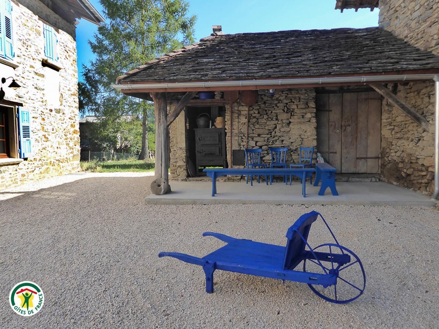 A rustic stone building with a covered patio area, outdoor seating, and a blue wheelbarrow.