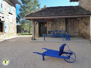 A rustic stone building with a covered patio area, outdoor seating, and a blue wheelbarrow. - Adal-Berht - SAINT MAXIMIN (SAINT MAXIMIN)