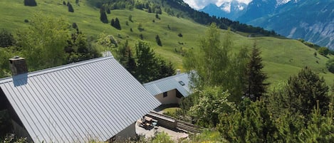 Een huis met schuin dak, omgeven door een weelderig groen landschap en bergen op de achtergrond.