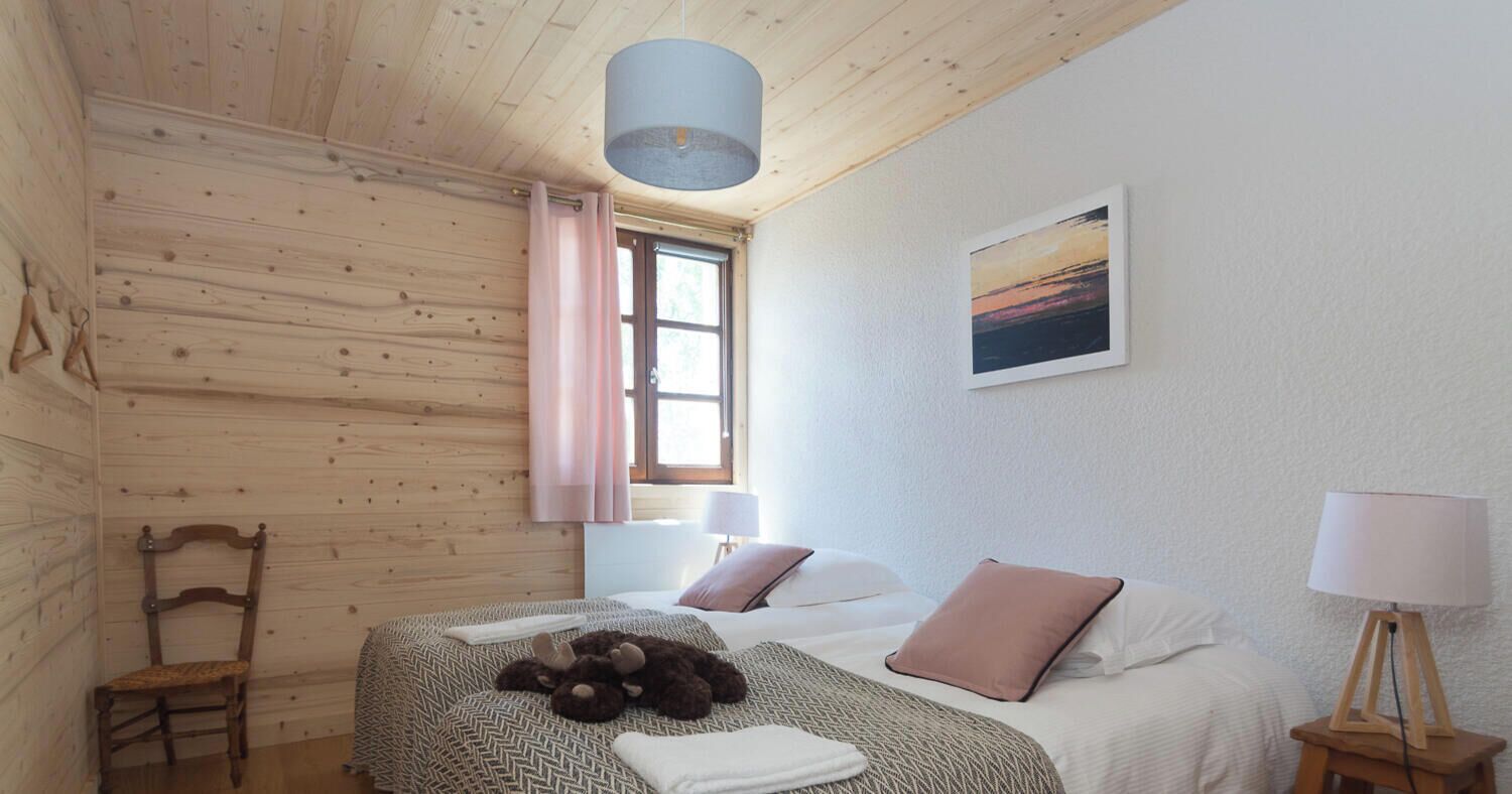A room with wooden walls, a bed with two pillows, a chair, a window with curtains, a lamp, and a painting on the wall.